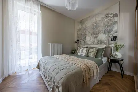 Przytulna sypialnia z dużym łóżkiem w neutralnych odcieniach, ozdobiona tropikalną tapetą nad zagłówkiem, naturalnymi dodatkami i tekstyliami.