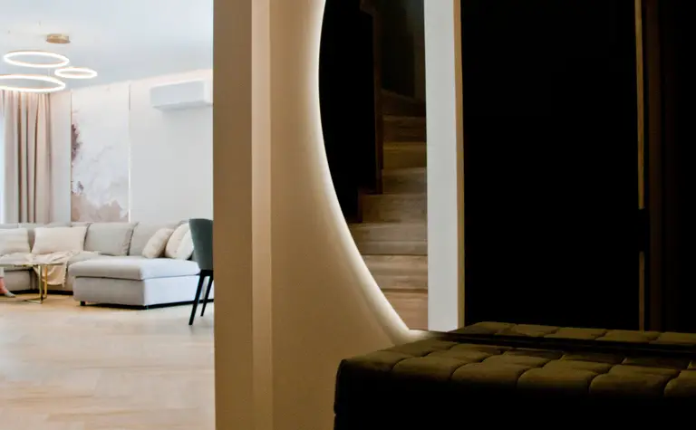 Nowoczesny korytarz z podświetlanym, okrągłym przejściem do salonu, w którym znajduje się jasna sofa i designerskie lampy wiszące. Schody i elegancka ławka w tle dodają wnętrzu stylu.