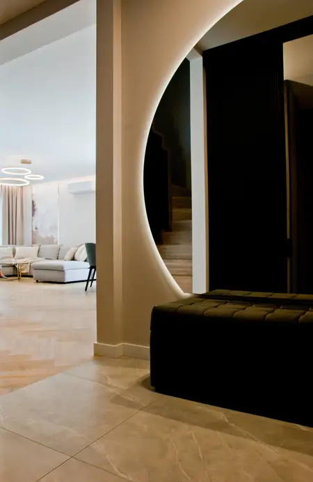 Nowoczesny korytarz z podświetlanym, okrągłym przejściem do salonu, w którym znajduje się jasna sofa i designerskie lampy wiszące. Schody i elegancka ławka w tle dodają wnętrzu stylu.