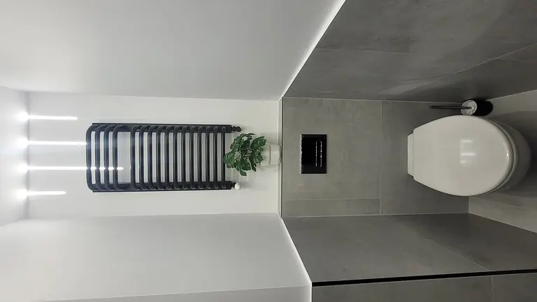 Toaleta z szarymi płytkami, białą toaletą i rośliną w doniczce, minimalistyczny design.