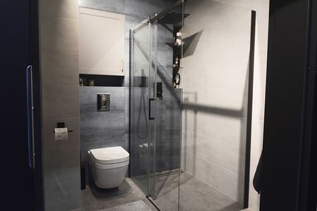 Wnętrze łazienki z minimalistycznym designem, szarymi płytkami i przezroczystą kabiną prysznicową.
