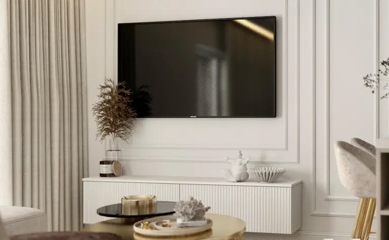 Stylowy salon z telewizorem na ścianie, białymi panelami dekoracyjnymi, złotym stolikiem kawowym i delikatnymi dodatkami w jasnych tonacjach