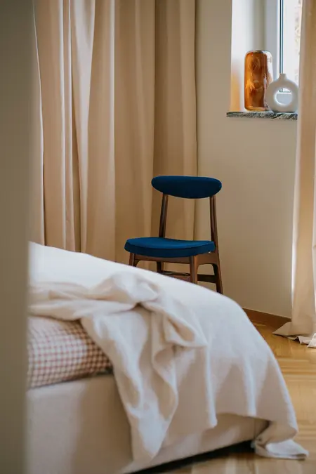 Wnętrze sypialni z beżowymi zasłonami i niebieskim krzesłem w stylu vintage i soft minimalizm.