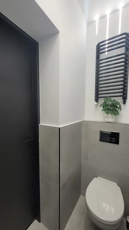 Toaleta w stylu minimalistycznym z szarymi płytkami, czarnymi akcentami i rośliną.