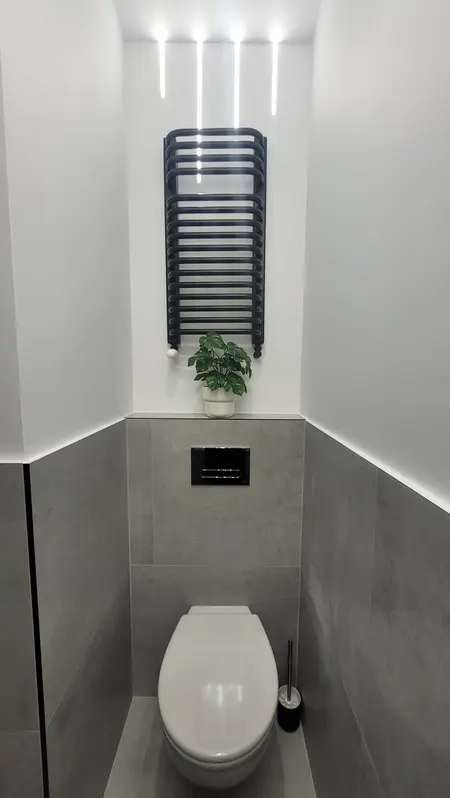 Toaleta z szarymi płytkami, białą toaletą i rośliną w doniczce, minimalistyczny design.