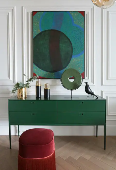Wnętrze z zieloną komodą, czerwonym taboretem oraz dekoracjami w stylu minimaluxe i quiet luxury.