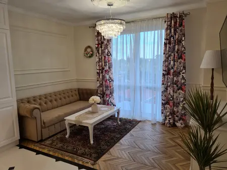 Tradycyjny salon z beżową sofą, kwiatowymi zasłonami i drewnianą podłogą.