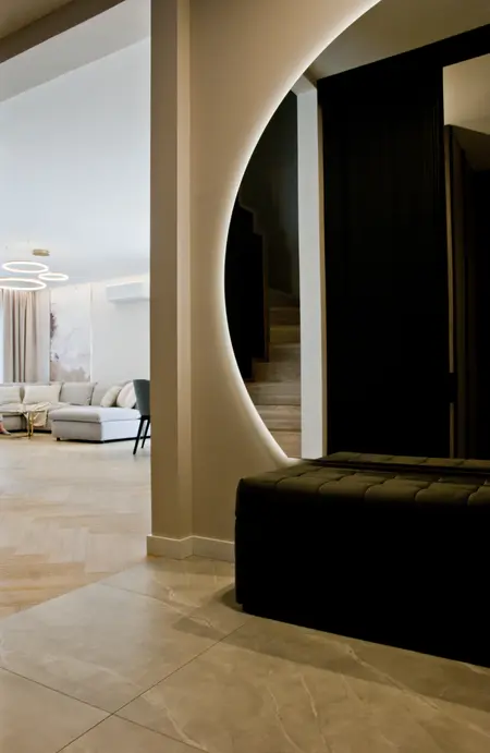 Nowoczesny korytarz z podświetlanym, okrągłym przejściem do salonu, w którym znajduje się jasna sofa i designerskie lampy wiszące. Schody i elegancka ławka w tle dodają wnętrzu stylu.