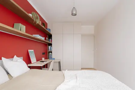 Sypialnia minimalistyczna z czerwoną ścianą, drewnianymi półkami i nowoczesnym biurkiem.