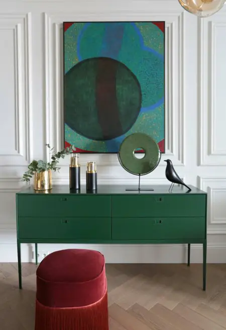 Wnętrze z zieloną komodą, czerwonym taboretem oraz dekoracjami w stylu minimaluxe i quiet luxury.
